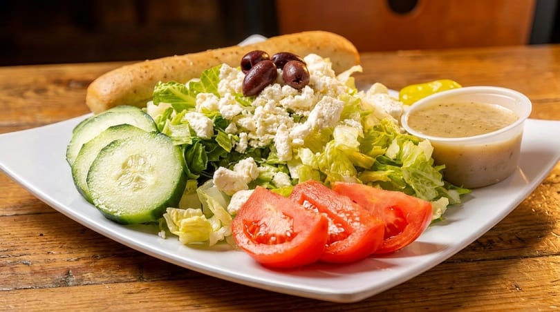 A fresh Greek salad with lettuce, feta, olives, cucumbers, tomatoes, and a breadstick.