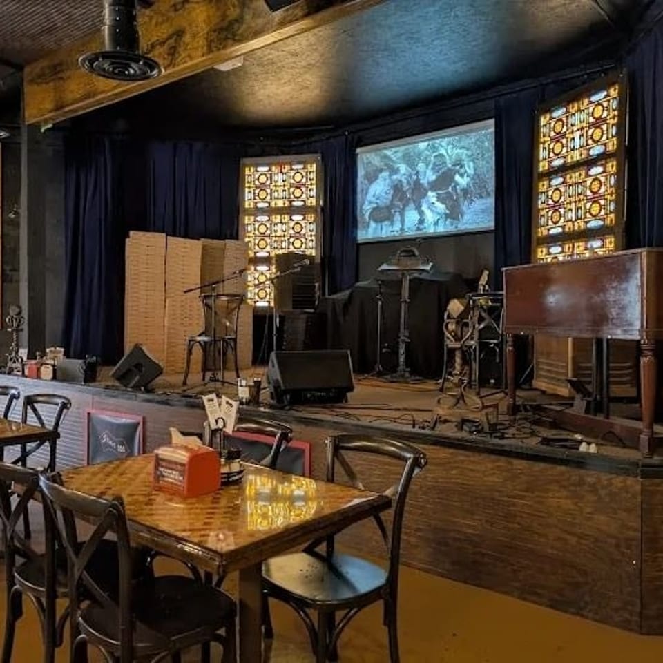 Restaurant dining area facing a stage with musical equipment, a projection screen, and stained glass panels.