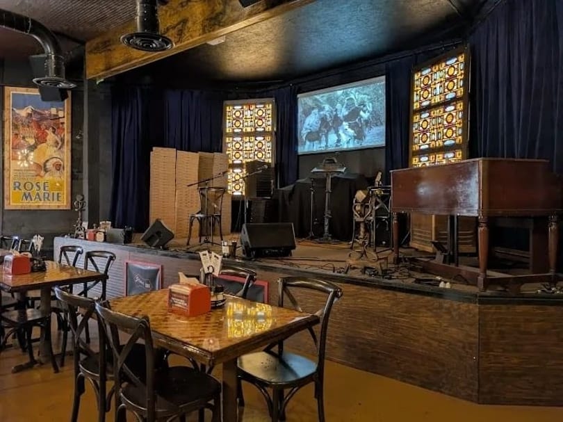 Restaurant dining area facing a stage with musical equipment, a projection screen, and stained glass panels.