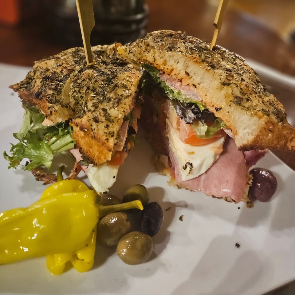Focaccia sandwich cut in half, revealing ham, mozzarella, lettuce, and tomato, served with olives and a yellow pepper.