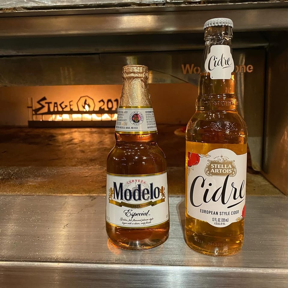 Modelo Especial beer and Stella Artois Cidre bottles on a counter in front of a lit pizza oven.