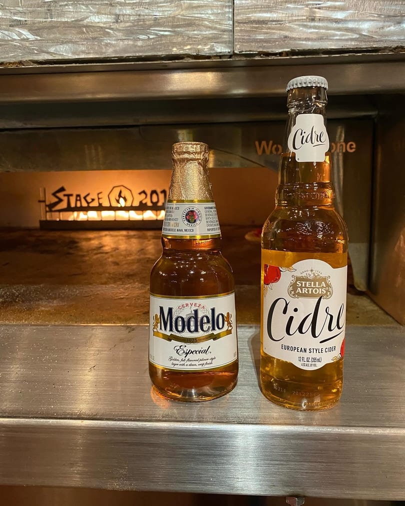 Modelo Especial beer and Stella Artois Cidre bottles on a counter in front of a lit pizza oven.