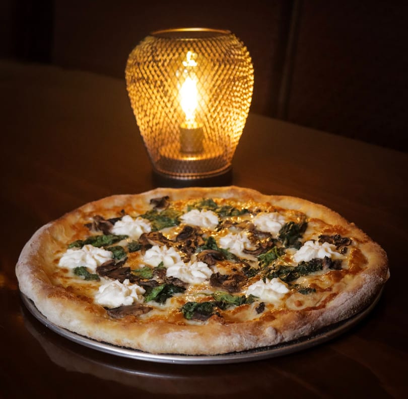 Pizza with ricotta, spinach, and mushrooms on a dark table, lit by a warm lamp.