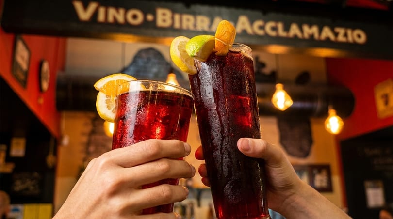 Two hands clinking tall glasses of red sangria garnished with citrus slices, under a "Vino Birra" sign.