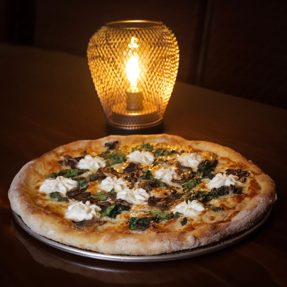 Pizza with ricotta, spinach, and mushrooms on a dark table, illuminated by a warm lamp.