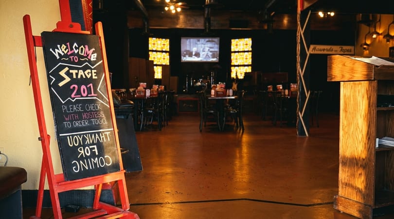A welcome sign for Stage 201 stands in a dimly lit restaurant with tables and a stage.
