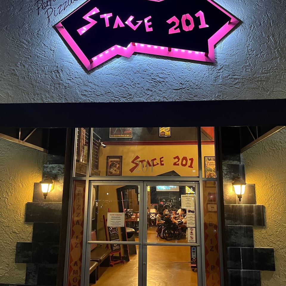 Stage 201 restaurant exterior at night, featuring a glowing pink neon sign and visible diners.