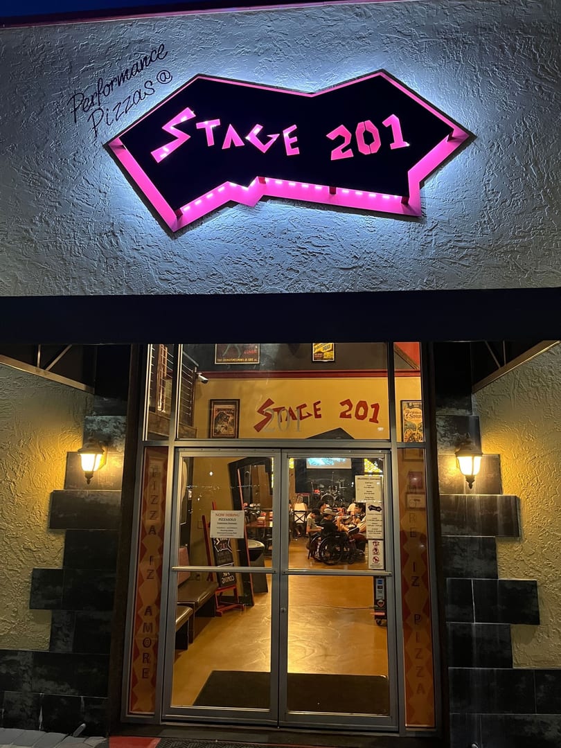 Stage 201 restaurant exterior at night, featuring a glowing pink neon sign and visible diners.