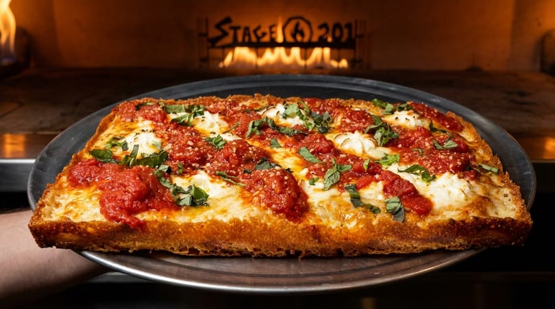 A rectangular Detroit-style pizza with red sauce, white cheese, and basil, held in front of a pizza oven.