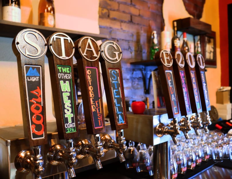Close-up of multiple beer taps with various brand names like Coors, West, Oktoberfest, and Stella.